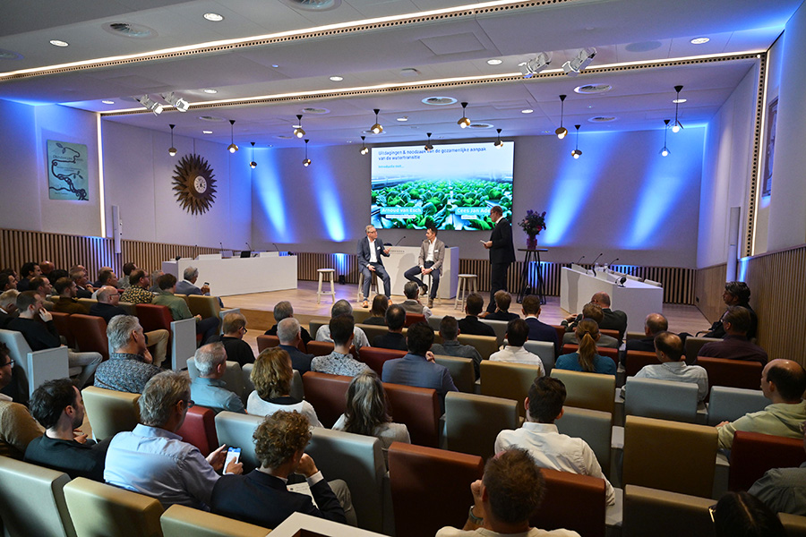 Grote zaal vol mensen en twee mannen op het podium zijn in gesprek.