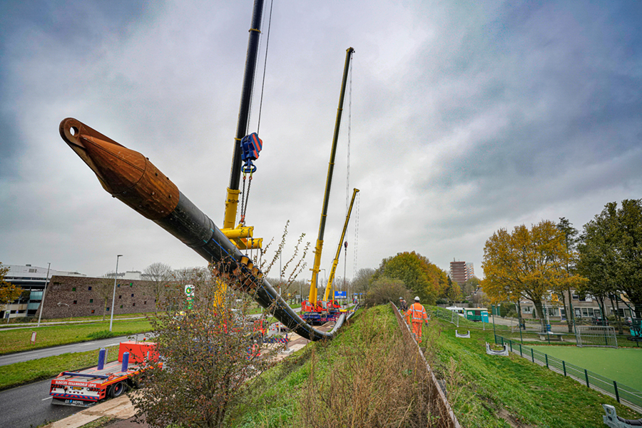 Grote transportleiding hangt aan drie kranen in de lucht, langs een weg.